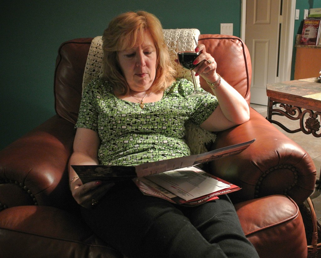a woman sitting in a chair reading a magazine and drinking wine